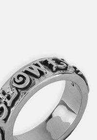 Srebrni prsten s uzdignutim, stiliziranim natpisima i zakrivljenim linijama. Glatka, polirana površina, okrugla traka i minimalistički dizajn.