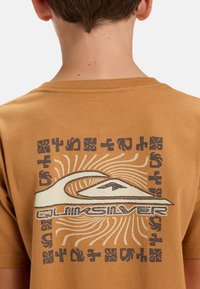 Camiseta de algodón marrón con un diseño gráfico que presenta una ola, montañas, rayos de sol y patrones tribales en colores negro y crema en la parte posterior.
