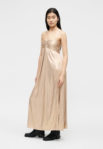 Γυναίκα με μακριά σκοτεινά μαλλιά φοράει μακρύ μπεζ σατέν φόρεμα με λεπτές τιράντες, συνδυασμένο με μαύρα μποτάκια αστραγάλου με αλυσίδες, στέκεται μπροστά σε γκρι φόντο.