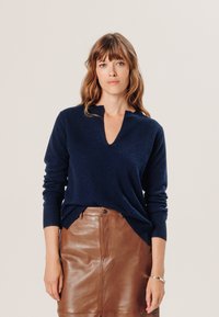 Marineblaues, langärmliges Strickpullover mit V-Ausschnitt und gerippten Bündchen, kombiniert mit einem braunen Lederrock. Weiche Textur und lockere Passform.