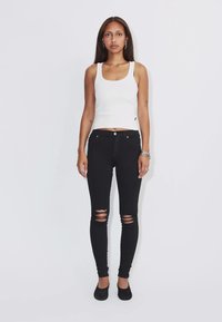 Donna in piedi dritta con jeans skinny neri strappati, top bianco senza maniche, scarpe nere e gioielli d’argento su sfondo bianco uniforme.
