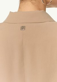 Beżowy kawałek odzieży z kołnierzykiem o gładkiej fakturze, z małym srebrnym logo z tyłu oraz pionowymi detalami szwów.
