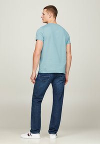 Camiseta de manga corta de color azul claro, con cuello redondo, combinada con jeans de mezclilla de color azul oscuro. El modelo está de espaldas a la cámara, con zapatillas blancas visibles.