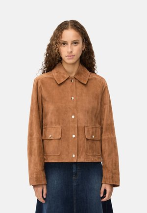 Giovane donna con capelli ricci che indossa una giacca in suede marrone con tasche frontali e bottoni argento, abbinata a una gonna di denim blu scuro.