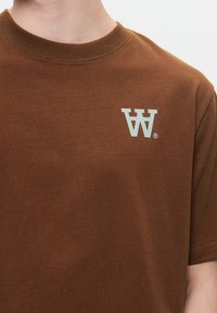 Brown cotton t-shirt featuring a round neckline and short sleeves. The left chest displays a light blue "W" logo, indicating branding.