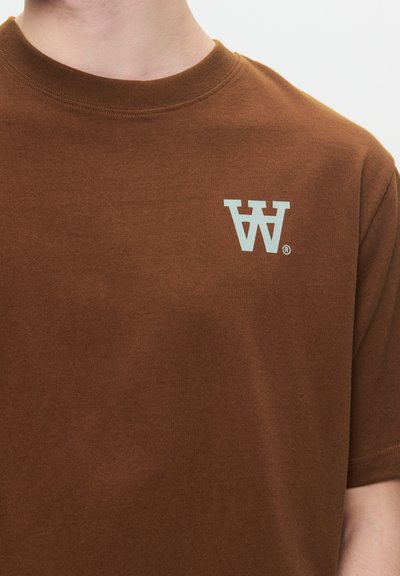 T-shirt in cotone marrone con scollo rotondo e maniche corte. Sul lato sinistro del petto è stampato un logo "W" azzurro chiaro, che indica il marchio.