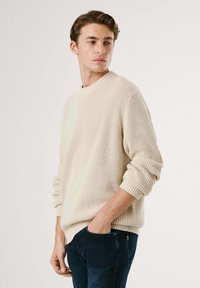 Young man with light beige knit sweater and dark jeans, standing with one hand in pocket, looking to side against plain background.