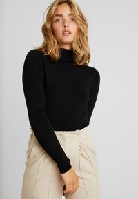 Pull en col roulé noir à manches longues, fabriqué dans un matériau doux. Porté avec un pantalon taille haute beige présentant des détails plissés.