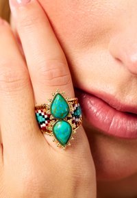 Gold ring featuring two teardrop turquoise stones, with a colorful beaded band in red, white, and blue. Smooth texture, intricate detailing.