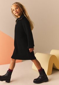 Abito nero lavorato a maglia con colletto e texture a coste, indossato con stivali alla caviglia con suola spessa e calze bordeaux. Sfondo neutro.