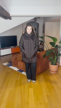 Črna podložena parka z visokim ovratnikom, sprednjim zadrgo, bočnimi žepi in elastičnimi manšetami. Nosi se čez temne hlače s srebrnimi supergami. Notranji prostor.