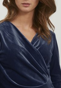 Navyblaues Samt-Wickeloberteil mit tiefem V-Ausschnitt und sanften Drapierungen. Der Stoff hat eine glatte Textur und einen leichten Glanz, der den eleganten Look unterstreicht.