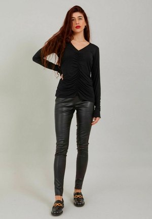 Frau mit langen roten Haaren, die ein gerafftes schwarzes Langarmshirt, schwarze Lederhosen und schwarze Loafers mit goldenen Kettendetails trägt, steht selbstbewusst.