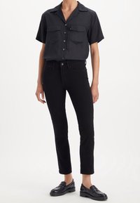 Camisa negra de manga corta con botones y dos bolsillos en el pecho, combinada con unos vaqueros negros ajustados y unos mocasines negros brillantes. Textura suave en todo.