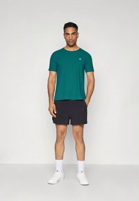 Kortærmet t-shirt i teal kombineret med sorte shorts. Stoffet virker glat; designet er enkelt med et lille logo på brystet. Båret med hvide sneakers.