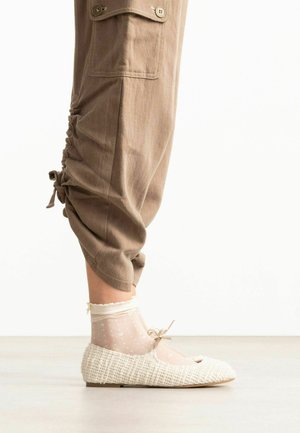 Been in cropped bruine broek, doorschijnende witte enkelsokken met stippen en geweven witte ballerina’s met kleine strikjes op een lichte vloer.