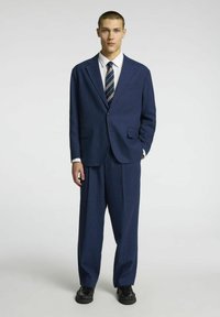Veste bleu marine en tissu texturé, dotée d'un revers cranté et de deux poches avant, accompagnée d'une cravate à motif et de chaussures noires.