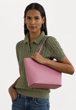 Rosa læder tote taske med en struktureret form, glat tekstur og kontrasterende sorte detaljer. Modellen holder tasken og bærer en grøn tekstureret sweater og jeans.