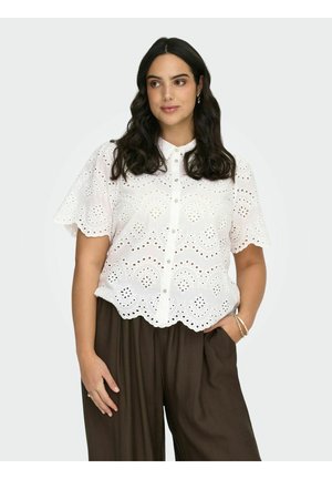 Kvinde med langt mørkt hår iført hvid bluse med hulmønster og korte ærmer samt løstsiddende brune bukser, poser med den ene hånd i lommen mod en hvid baggrund.
