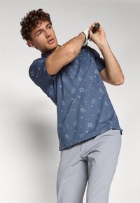 Blå kortermet polo skjorte med subtile mønstre; brukt med lysegrå golfbukser. Modellen svinger en golfkølle, og viser frem stoff og passform.