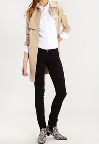 Abrigo trench beige sobre una camisa blanca, combinado con jeans negros ajustados y botas de tobillo en blanco y negro con estampado de animal.
