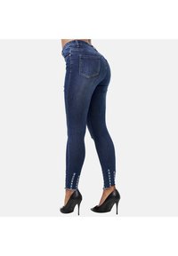 Jeans ajustados de mezclilla azul oscuro, con detalles rasgados en el dobladillo. Combinados con tacones altos negros, que destacan una silueta ceñida.