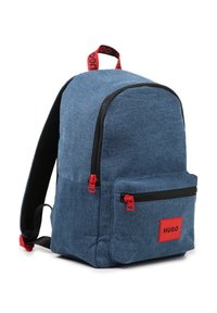 Denim backpack in dark blue with red zippers and logo patch. Features a large main compartment and a front zip pocket for storage.