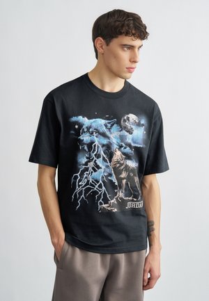 Young man wearing black T-shirt with graphic of howling wolf, snarling wolf with lightning, and moon, paired with gray pants.