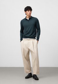 Dunkelgrünes, gestricktes Polohemd mit langen Ärmeln und Kragen, kombiniert mit beigen, weit geschnittenen Hosen. Schwarze Lederschuhe runden das Outfit ab.
