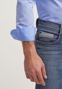 Mands venstre arm med optrukket ternet blå skjortesærme og hånd hvilende ved siden af blå denim-jeanslomme synlig.