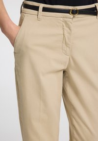 Béžové kalhoty vyrobené z bavlněné směsi, s elegantním designem, bočními kapsami a černým koženým páskem se zlatou sponou.