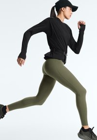Femme en haut noir à manches longues, casquette noire, leggings verts et baskets noires courant sur un fond blanc.