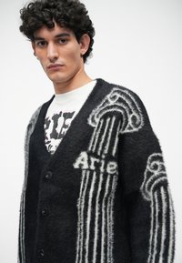 Cárdigan negro con textura y patrón de columnas blancas, con el texto "Arie". Cuenta con un escote en V, botones y un ajuste holgado. Superpuesto sobre una camiseta gráfica.