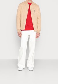 Uomo che indossa una giacca a trapuntatura beige sopra una camicia rossa, pantaloni bianchi a gamba larga e sneakers bianche, in piedi contro uno sfondo bianco semplice.