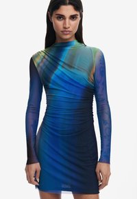 Tightfittad långärmad klänning i gradientblå och grön tyg. Har en ruchad design och genomskinliga ärmar med subtila mönsterdetaljer.
