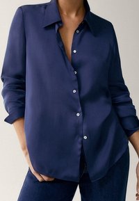 Camicia blu navy con bottoni, colletto classico, maniche lunghe e vestibilità comoda. La texture liscia e i bottoni bianchi creano un contrasto.