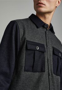 Camicia button-up con corpo grigio texturizzato e maniche blu navy, dotata di due tasche frontali e bottoni neri. Design del colletto distintivo.