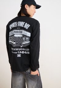 Black long-sleeve t-shirt with white graphic print featuring "SPORTS STORE DAY" and storefront design. Worn with gray cargo pants.