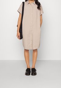 Robe chemise beige à manches courtes et à boutonnage sur le devant, réalisée en un tissu léger. Associée à des sabots noirs et à un sac à bandoulière noir texturé.