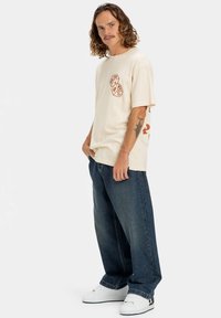 Beige oversized t-skjorte med grafiske detaljer, paret med mørkeblå baggy jeans og hvite sneakers. Korte ermer og myk tekstur.