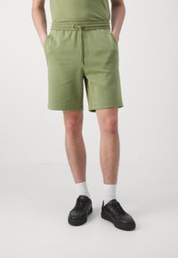 Bermudas de algodón verde claro con cintura elástica y cordón, que cuentan con bolsillos laterales y un corte relajado. Combinadas con zapatillas negras.