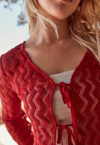 Cardigan all'uncinetto rosso con motivo a zigzag, maniche lunghe, apertura frontale e nastri rossi. Indossato sopra un top bianco. Tessuto lavorato a maglia.