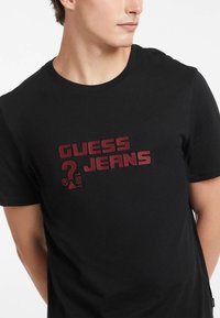 T-shirt en coton noir avec une impression graphique rouge qui indique "GUESS JEANS 1981". Col rond et manches courtes. Texture douce.
