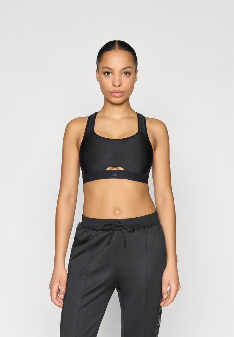 Μαύρο αθλητικό σουτιέν με στρογγυλή λαιμόκοψη και άνοιγμα σε σχήμα κλειδαρότρυπας, κατασκευασμένο από μονόχρωμο ύφασμα και πλατιά τιράντες. Συνδυάζεται με μαύρο στενό παντελόνι jogging με ελαστική μέση.