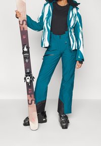 Teal ski pants with zip pockets and black accents paired with a white and teal patterned puffer jacket. A black t-shirt and skis complete the outfit.