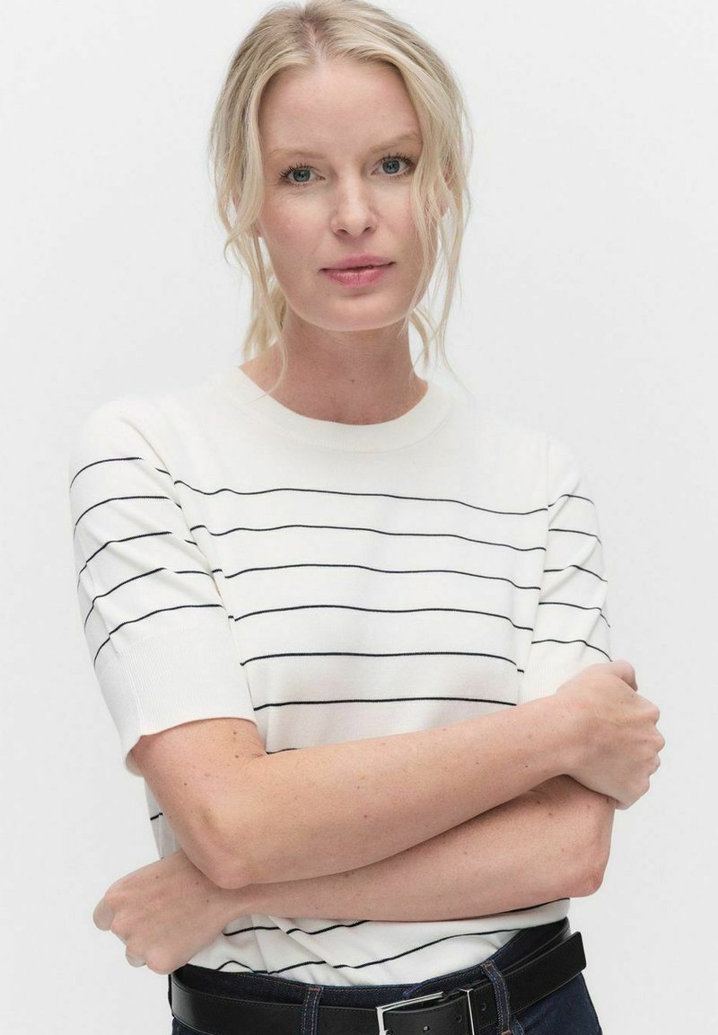 Hvid kortærmet sweater med vandrette marineblå striber og rund halsudskæring, parret med mørke jeans og et sort bælte.