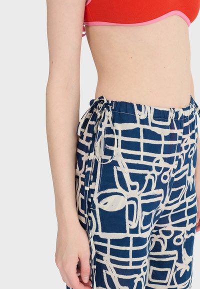 Widok z boku osoby noszącej czerwony krótki top i niebieskie spodnie z białą abstrakcyjną grafiką oraz wiązanym paskiem na bok.