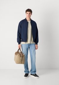 Veste bomber marine, sweat-shirt beige, jean bleu clair, baskets noires et blanches, tenant un sac de sport en suède beige.