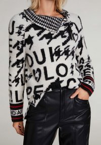 Pull en maille blanc et noir avec texte et symboles audacieux, col pied-de-poule et poignets rayés, assorti à un pantalon en fausse cuir noir.