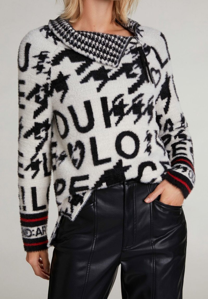 Pull en maille blanc et noir avec texte et symboles audacieux, col pied-de-poule et poignets rayés, assorti à un pantalon en fausse cuir noir.